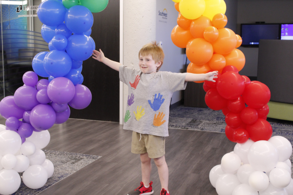 A pre-k student smiles underneath a rainbow balloon arch.