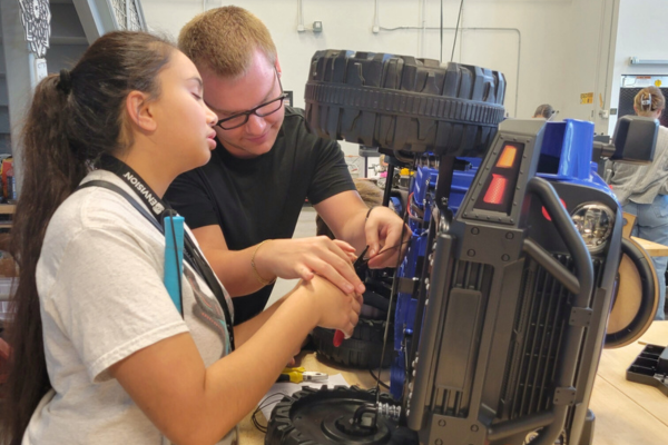 A teenage girl who is visually impaired works on an electric car, holding her cane.