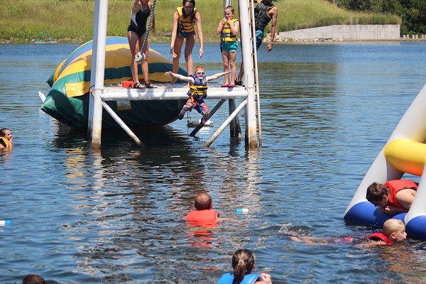 Envision campers in the water with a big ledge in the middle. A few people are standing on the ledge watching a young boy jump in the water with his arms and legs out and he has a big smile on his face. 